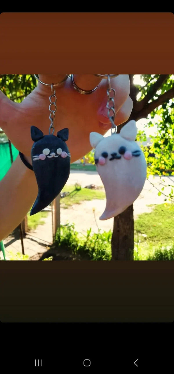 Llaveros kawaii de gatos fantasma blanco y negro con mejillas sonrosadas, colgando en un entorno natural