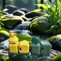 Yellow rubber duck and green frog keychain toys on mossy rocks by a flowing stream